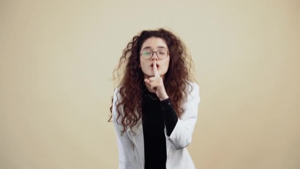 The serious young woman with curly hair says to keep her finger on her lips, leaning forward. Young hipster in gray jacket and white shirt, with glasses posing isolated on beige background in the - Footage, Video