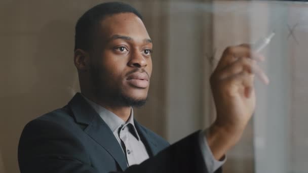 Портрет афро-американского бизнесмена, стоящего в офисе и пишущего маркером на прозрачной стеклянной стене, принимая к сведению уравнение, развивающее стратегию проекта, рассматривает финансовую прибыль улыбающейся - Кадры, видео