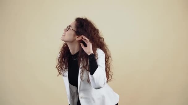 Curious young woman with curly hair, with her hand to her ear, listened to gossip leaning forward. Young hipster in gray jacket and white shirt, with glasses posing isolated on beige background in the - Footage, Video