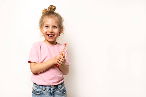 a small blonde girl smiles and holds a toothbrush on a white background, a place for text, the concept of caring for children's baby teeth and oral hygiene. - Photo, Image