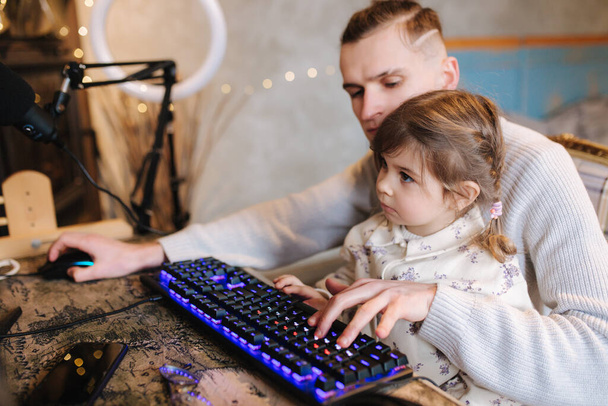 Younger sister sits on brothers knees and put finger on keyboard with neon light. Little girl help brother work on computer. Happy family - Photo, Image