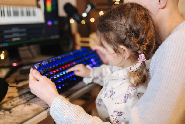 Младшая сестра сидит на коленях братьев и кладет палец на клавиатуру с неоновым светом. Девочка помогает брату работать за компьютером. Счастливая семья - Фото, изображение