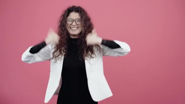 The young winner with curly hair, happy looks with both hands the gesture like smiling. Young hipster in white jacket and black shirt, with glasses posing isolated on pink background in the studio. - Footage, Video