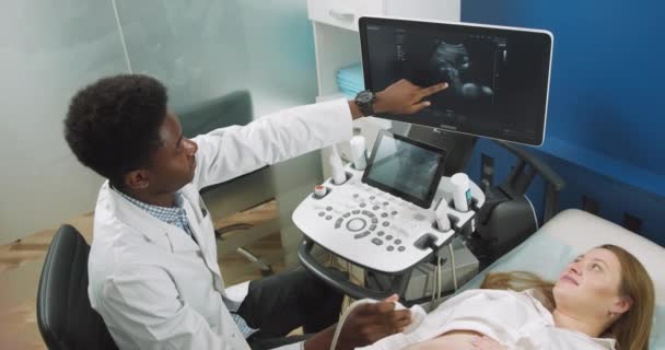 Έννοια υπερήχων. Κάτοψη της νεαρής Καυκάσιας εγκύου ξαπλωμένης στον καναπέ, ενώ ένας έμπειρος Αφροαμερικανός γιατρός κάνει υπέρηχο και δείχνει το μωρό στην οθόνη - Πλάνα, βίντεο