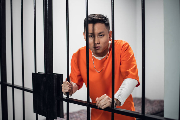 Convicted asian man in an orange robe, with a chain and bracelet and a tattoo on his face stands in a prison cell next to the bars - Photo, Image