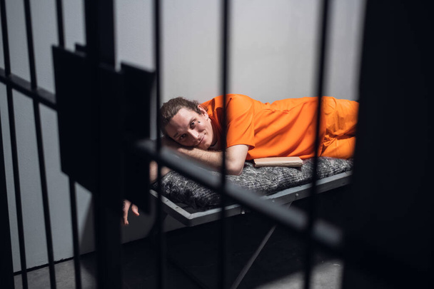 Τα φώτα σβήνουν στη φυλακή. Ένας κατάδικος με πορτοκαλί ρόμπα κάθεται σε μια κουκέτα σε ένα κελί με ένα βιβλίο.. - Φωτογραφία, εικόνα