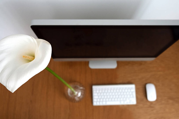 Werkplaats in een minimalistische stijl. Monitor, toetsenbord en muis op een houten tafel naast een thuisbloem en calla lelies. Kopieerruimte. . - Foto, afbeelding