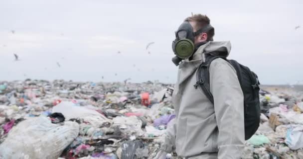 Symbolisch schot milieubescherming, man met gasmasker op stortplaats, rondkijken, camera bekijken, vuilnisbaan door de stortplaats op de achtergrond - Video