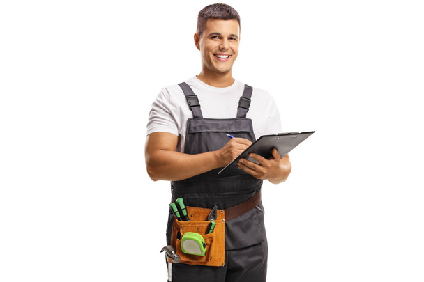 Smiling young repairman with a tool belt writing on a clipboard isolated on white background - Фото, зображення