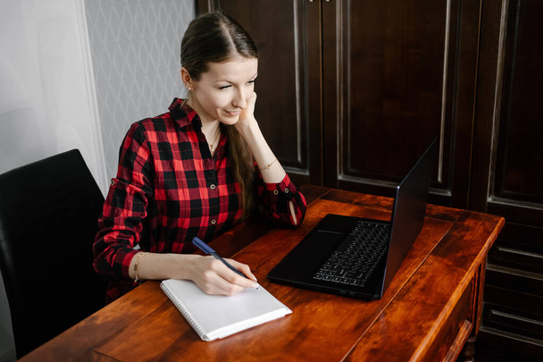 Happy european female student in  casual outfit, smiling while using laptop to learning online sitting at home interior. Self education in terms of quarantine. - 写真・画像