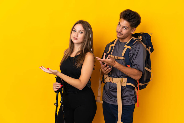 Young mountaineer couple with a big backpack isolated on yellow background extending hands to the side for inviting to come - 写真・画像