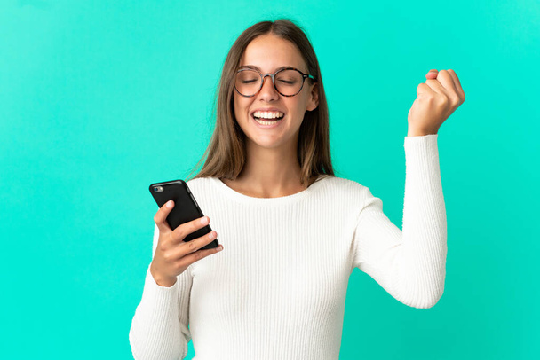 Mladá žena přes izolované modré pozadí pomocí mobilního telefonu a dělá vítězný gesto - Fotografie, Obrázek