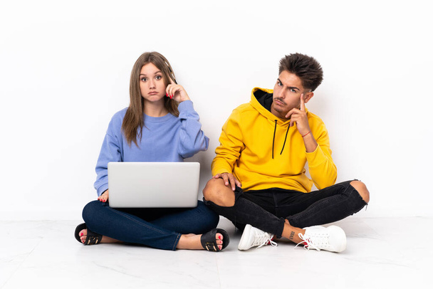 Giovane coppia con un computer portatile seduto sul pavimento isolato su sfondo bianco pensando un'idea - Foto, immagini