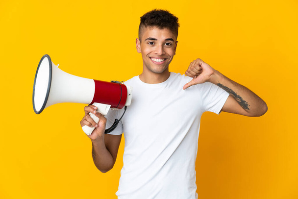 Young brazilian man isolated on yellow background holding a megaphone and proud and self-satisfied - Valokuva, kuva