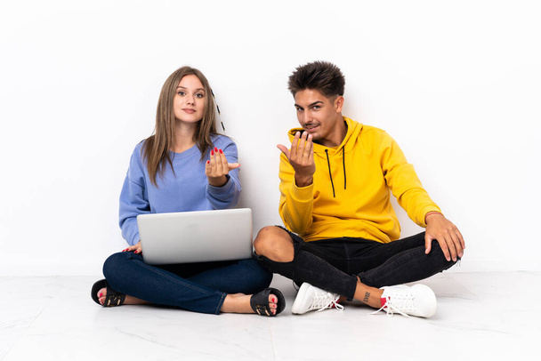 Jeune couple avec un ordinateur portable assis sur le sol isolé sur fond blanc invitant à venir avec la main. Heureux que tu sois venu - Photo, image
