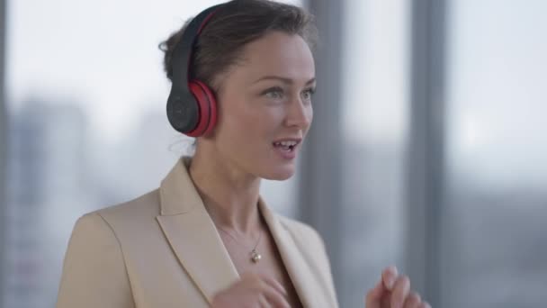 Positive slim beautiful woman in headphones enjoying music dancing at window. Portrait of happy relaxed Caucasian CEO employee having fun at break in office. Lifestyle and hobbies. - Imágenes, Vídeo