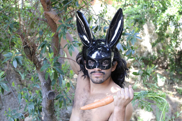 retrato de un hombre joven y guapo sin camisa con máscara de conejito negro sosteniendo zanahoria en el bosque - Foto, Imagen