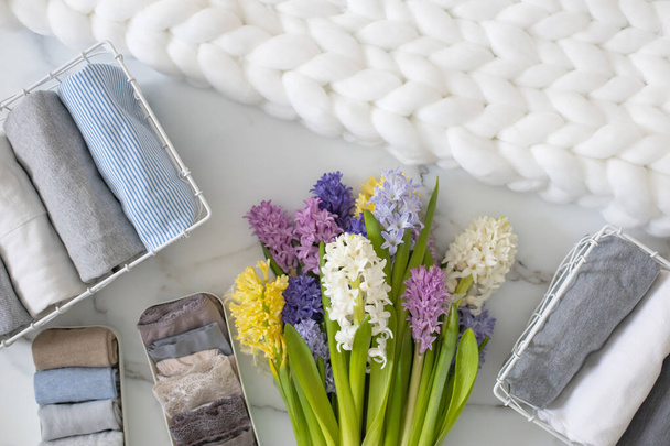 Stylish composition storage underwear organization Marie Kondo's method decorated by natural flowers. Top view contemporary folded container cases clothing on marble floor. White fluffy knitted plaid - Фото, зображення