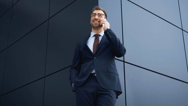 low angle view of happy businessman in suit and glasses talking on smartphone outside - Foto, imagen