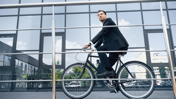 full length of businessman in suit riding bicycle near building with glass facade - 写真・画像