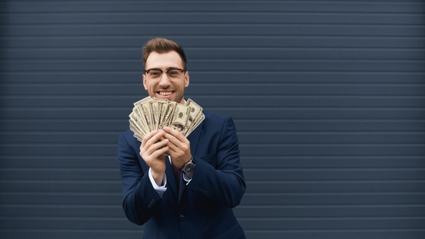 rich businessman in suit smiling while holding dollars  - Foto, afbeelding