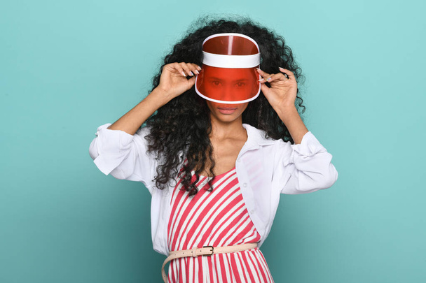 Trendikäs nuori musta tyttö yllään läpinäkyvä punainen huippu tai hattu kurkistaa visiiri kameran punainen ja valkoinen raidallinen toppi turkoosi studio tausta - Valokuva, kuva
