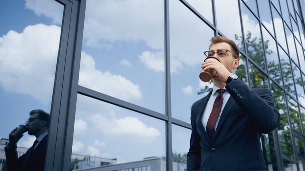 businessman in glasses drinking coffee to go near building with glass facade  - Photo, image