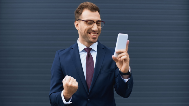 positive businessman in glasses and formal wear holding cellphone outside  - Fotó, kép