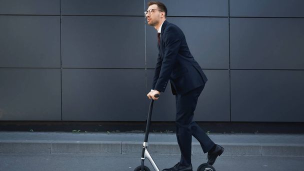 longitud completa de hombre de negocios en el desgaste formal y gafas de montar e-scooter cerca de la construcción  - Foto, Imagen