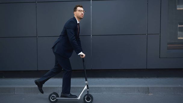 Geschäftsmann in Anzug und Brille fährt E-Scooter in der Nähe von Gebäude  - Foto, Bild