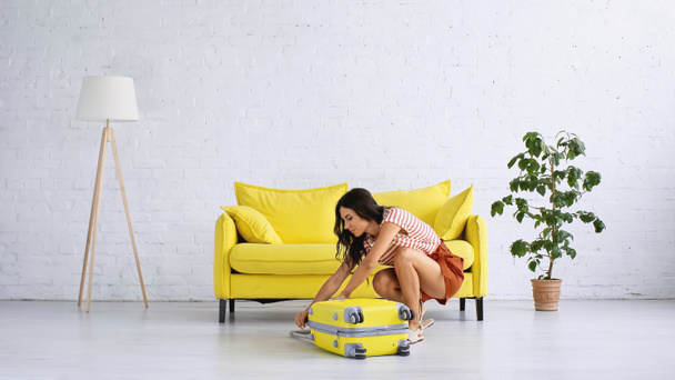 Brünette Frau schließt gelbe Reisetasche neben Couch im Wohnzimmer  - Foto, Bild