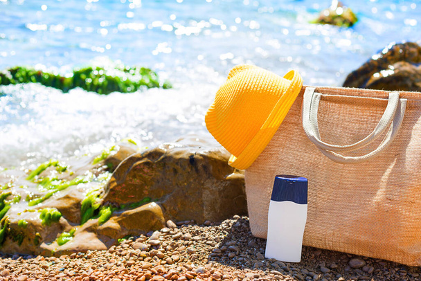 Beach bag with a white tube of sunscreen on a pebble beach near the sea. Travel, beach holiday at the resort, glasses, yellow hat. Protection of the skin from UV rays, suntan oil. Copy space, mock up. - Фото, зображення