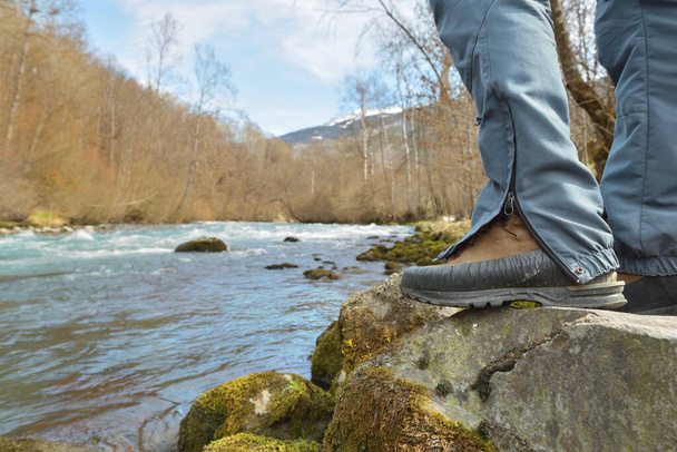 pies de un excursionista con zapatos de senderismo de pie sobre una roca sobre un río alpino que fluye en un valle  - Foto, Imagen