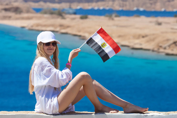 Chica rubia con ropa blanca sosteniendo la bandera de Egipto en el fondo del mar Rojo. Concepto de viajes y turismo. Enfoque selectivo. - Foto, Imagen