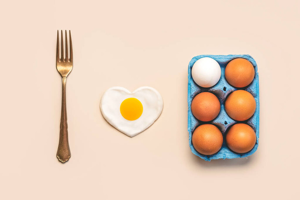 Κάτοψη αυγών κοτόπουλου σε ανοιχτό μπλε χάρτινο κουτί με vintage πιρούνι και τηγανητό αυγό σε σχήμα καρδιάς σε μπεζ φόντο - Φωτογραφία, εικόνα