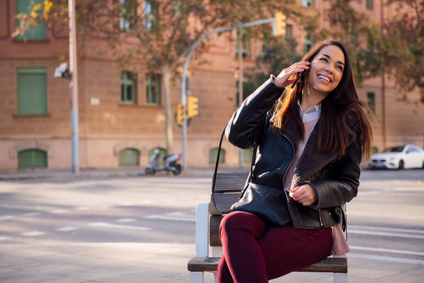 joyful young woman sitting on a street bench laughing while making a phone call, concept of communication and urban lifestyle, copyspace for text - Photo, image