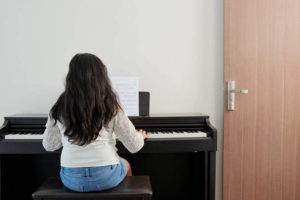 Kleines Mädchen lernt Symphonie und spielt zu Hause Klavier, Blick von hinten - Foto, Bild