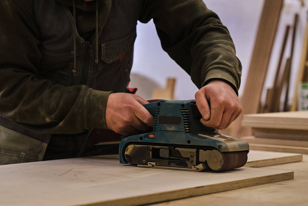 Κλείσε. Ξυλουργός σε εργαστήριο γυαλίζει ξύλινο πίνακα με ηλεκτρικό τροχιακό τριβείο. Έννοια ξυλουργικής και επίπλωσης. - Φωτογραφία, εικόνα