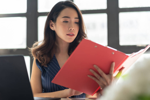 Portrait of Pretty young business woman holding and looking at red clipboard or document folder. Attractive Beautiful Asian female working in the office. - Foto, afbeelding