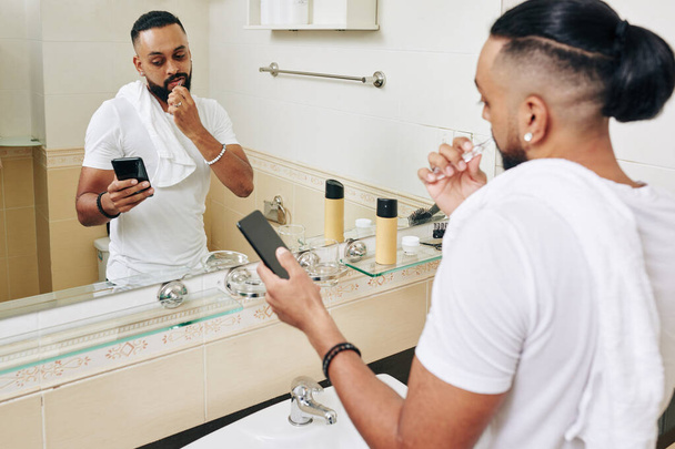 Серйозна людина чистить зуби у ванній кімнаті і читає новини на екрані смартфона
 - Фото, зображення