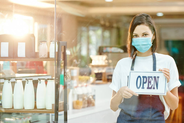 Portrait of young pretty woman in protective mask standing at shelf with milk and yogurt bottles in store and holding open sign - Photo, Image