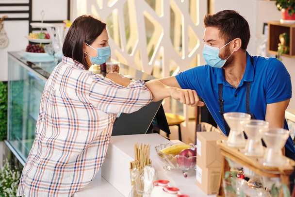 Coffeeshop barista και θηλυκό πελάτη σε ιατρικές μάσκες κάνοντας χτύπημα αγκώνα όταν χαιρετισμό ο ένας τον άλλον - Φωτογραφία, εικόνα