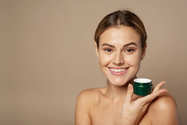 Portrait of a pretty young smiling woman with clean fresh skin in the studio on a beige background. Cute woman holding a jar of moisturizer in the studio. Skin care and hydration concept - Photo, Image