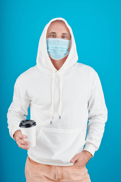 Porträt eines ernsthaften Mannes in Kapuzenpulli und medizinischer Maske, der eine Tasse Kaffee herausnimmt, isoliert auf blauem Grund - Foto, Bild