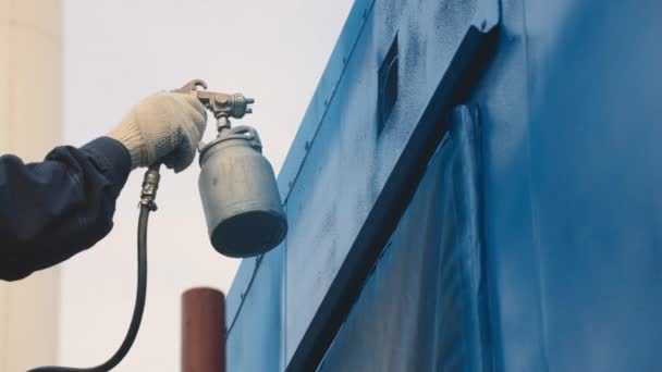 Un travailleur en salopette et un respirateur peignent la carrosserie d'une remorque cargo ou d'une voiture métallique en bleu.  - Séquence, vidéo