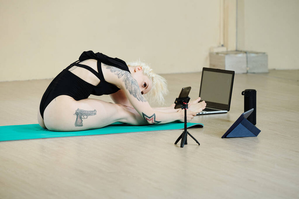 Ajuste mujer joven sentada en el suelo y inclinándose hacia adelante a sus piernas al filmar estiramiento entrenamiento - Foto, Imagen