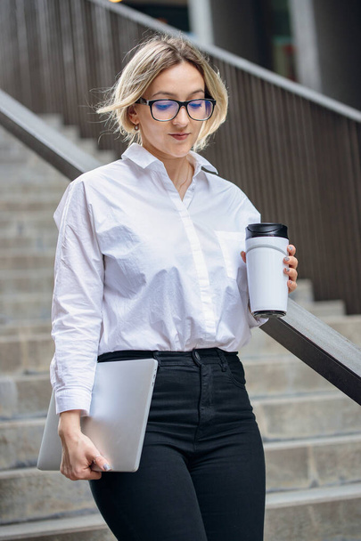 Liikenainen valkoisessa paidassa ja mustissa housuissa laskeutuu portaita. Nuori vaalea nainen kahvin kanssa uudelleenkäytettävässä kupissa, lasit ja kannettava tietokone. - Valokuva, kuva