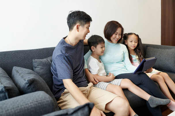 Happy Vietnamese family with two kids enjoying spending time together and watching animated video or blogs on digital tablet - Photo, image