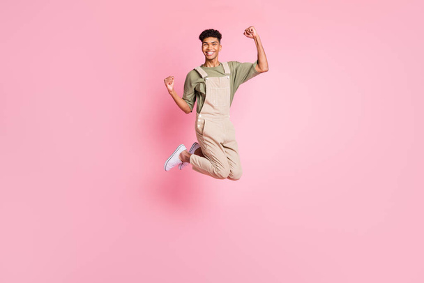 Full length body size photo of guy in overall jumping high gesturing like winner laughing isolated on pastel pink color background - 写真・画像