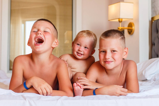 three cheerful laughing tanned beautiful children lie on the bed in a hotel room - Foto, imagen